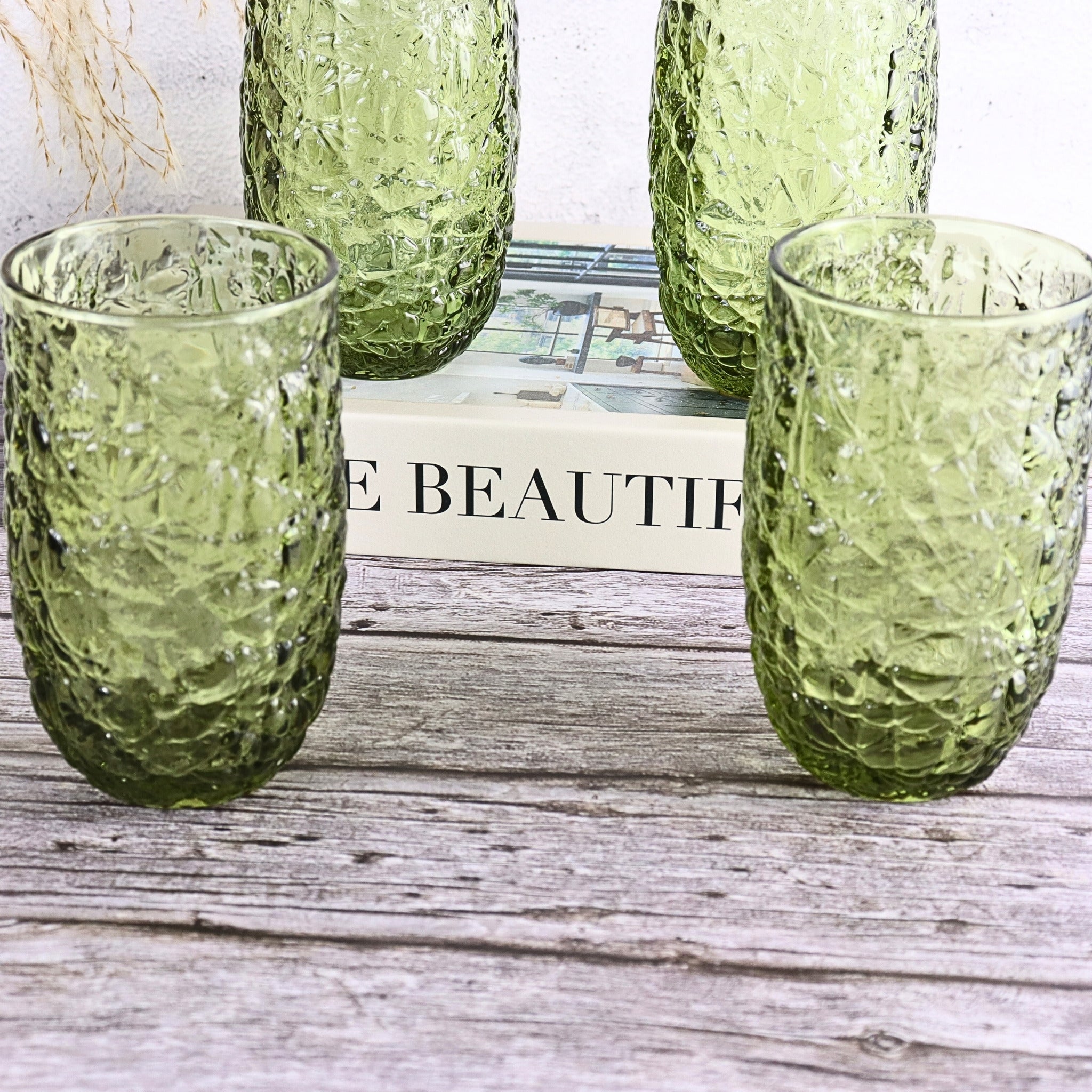 Four green textured glass tumblers on a wooden surface with a book in the background.