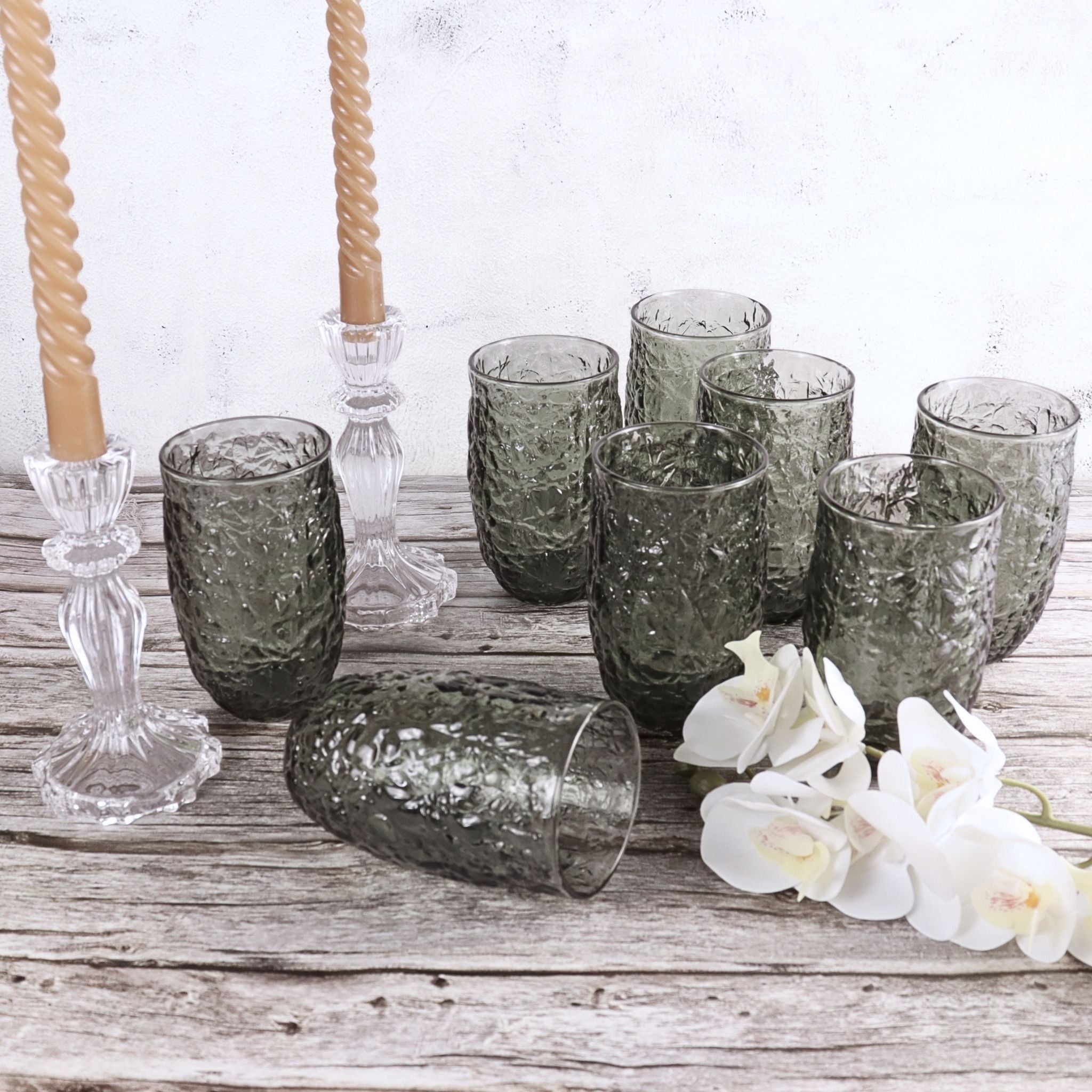 Set of smoked glass tumblers on a wooden surface with white flowers and candle holders.
