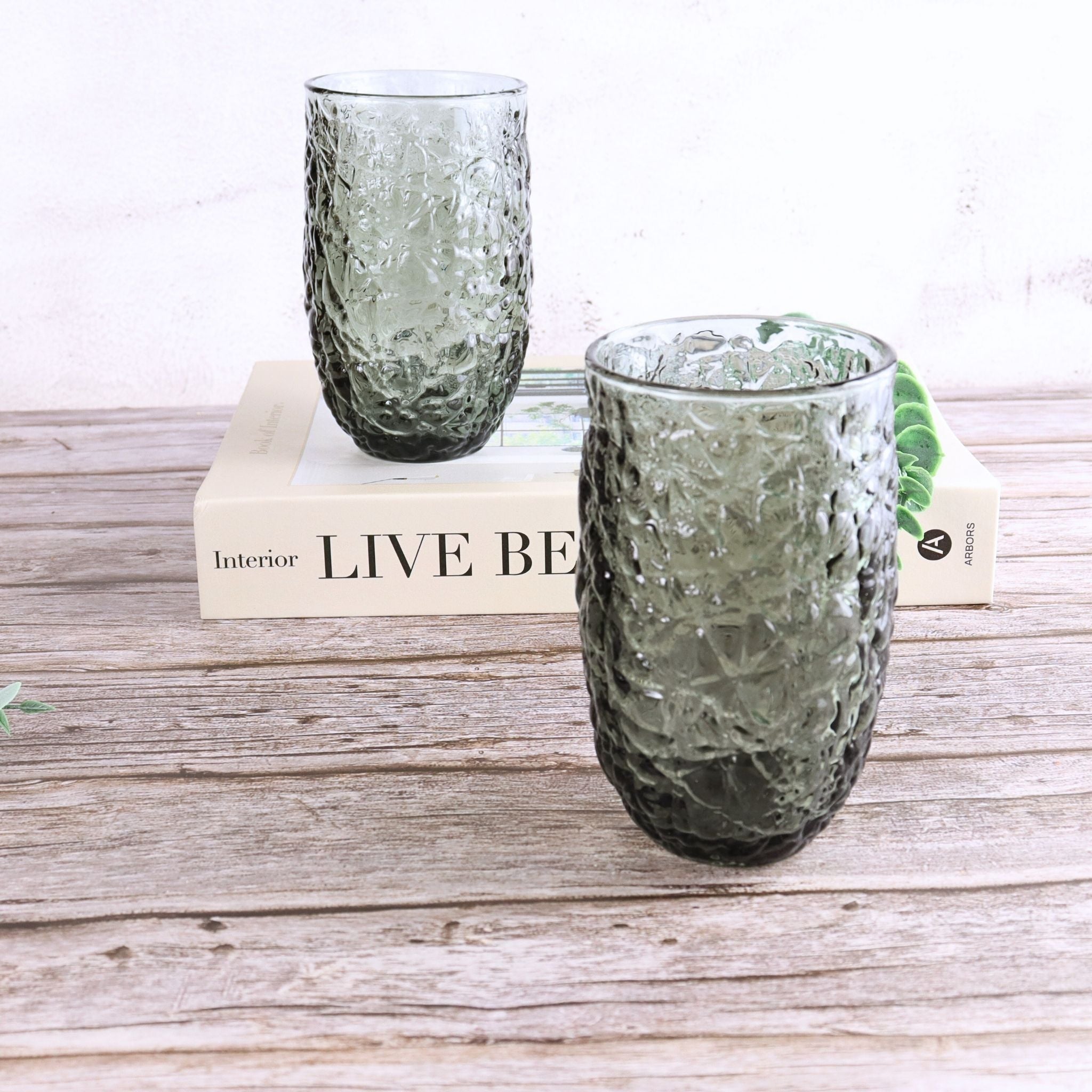 Two textured green glass tumblers on a wooden surface with a book underneath.