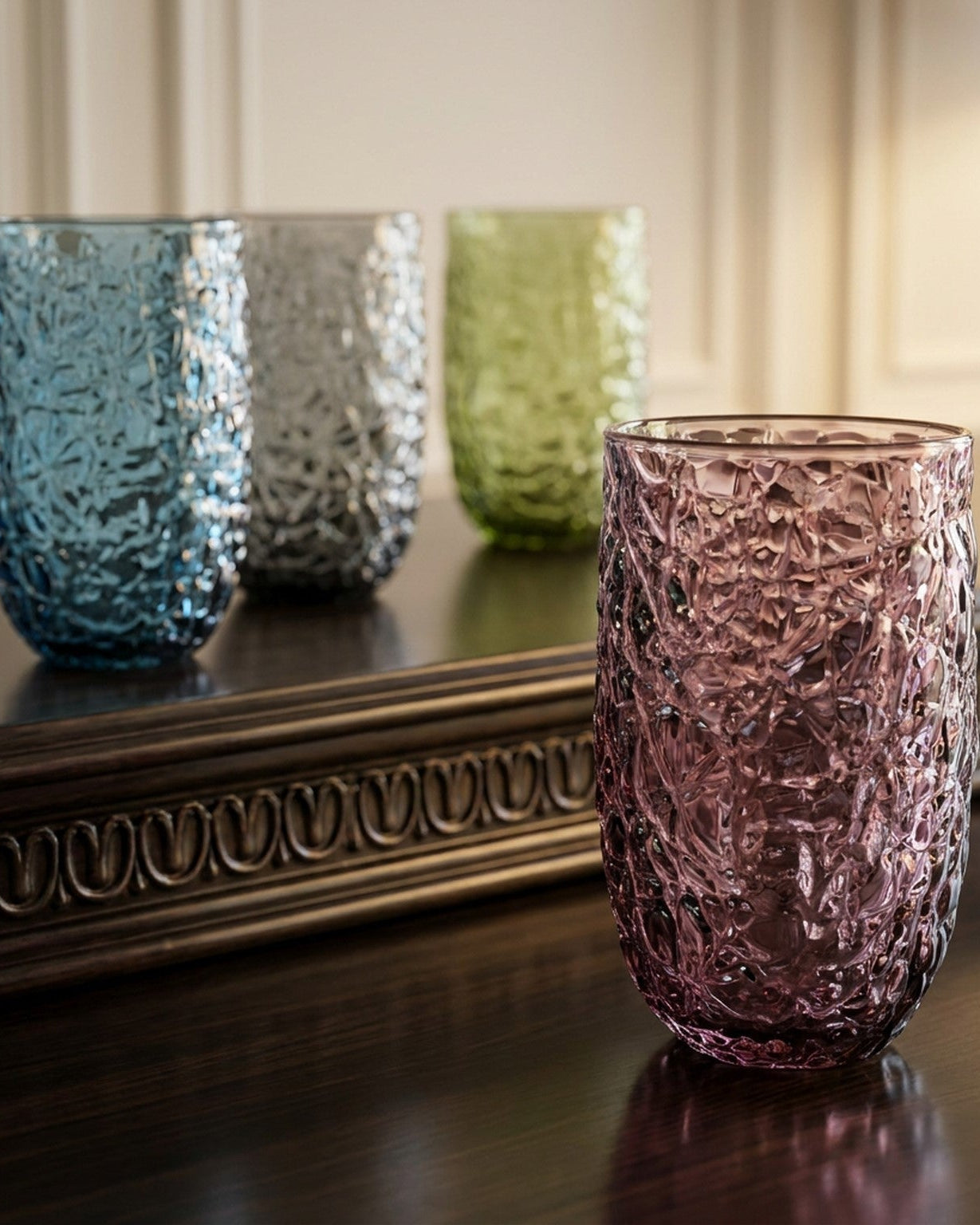 Four textured glass tumblers in pink, green, blue, and gray on a wooden surface.