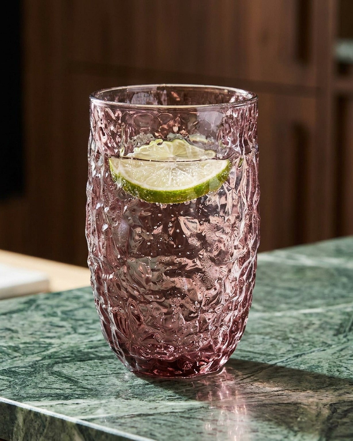 Two pink textured glasses on a wooden surface with a candle and greenery.