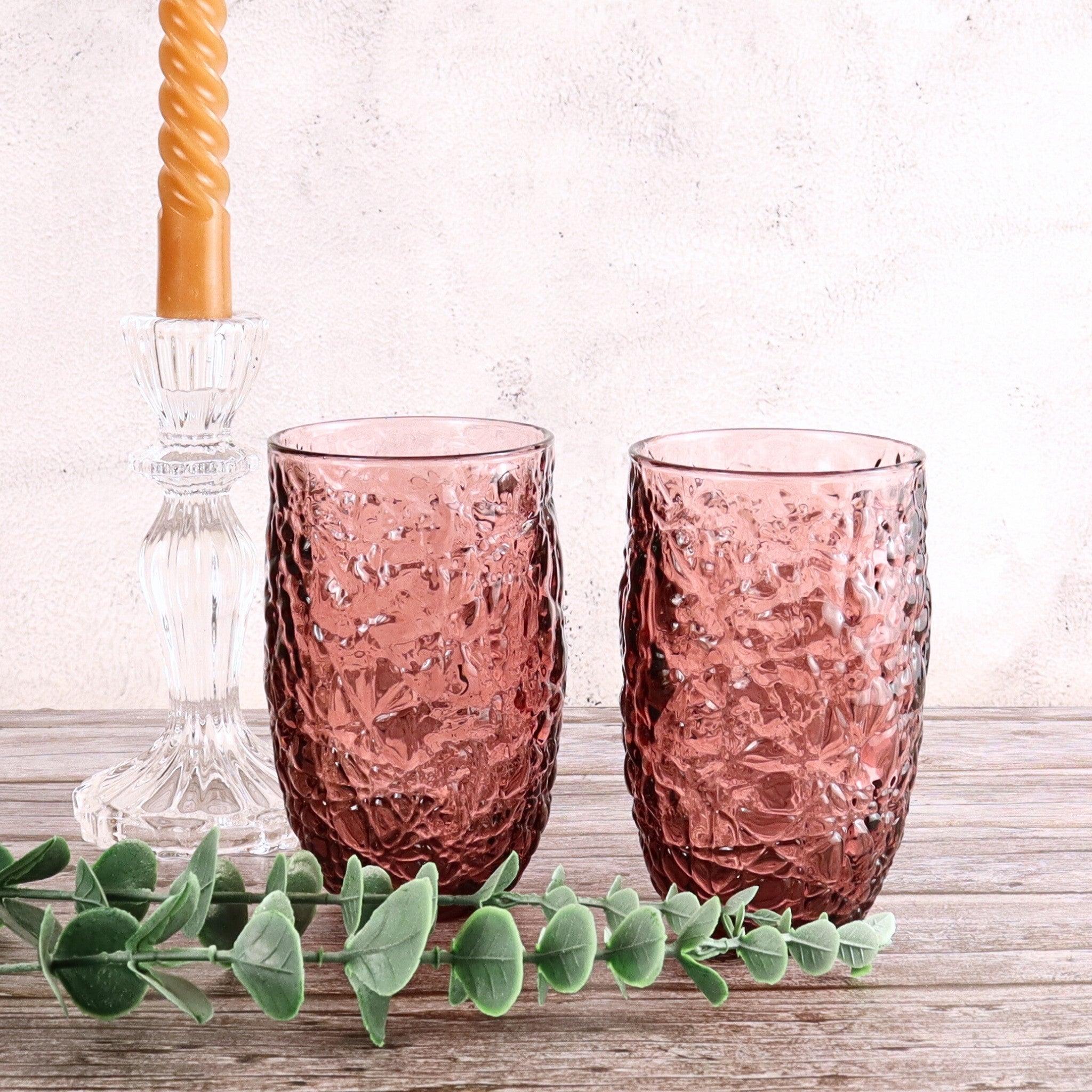 Two pink textured glasses on a wooden surface with a candle and greenery.