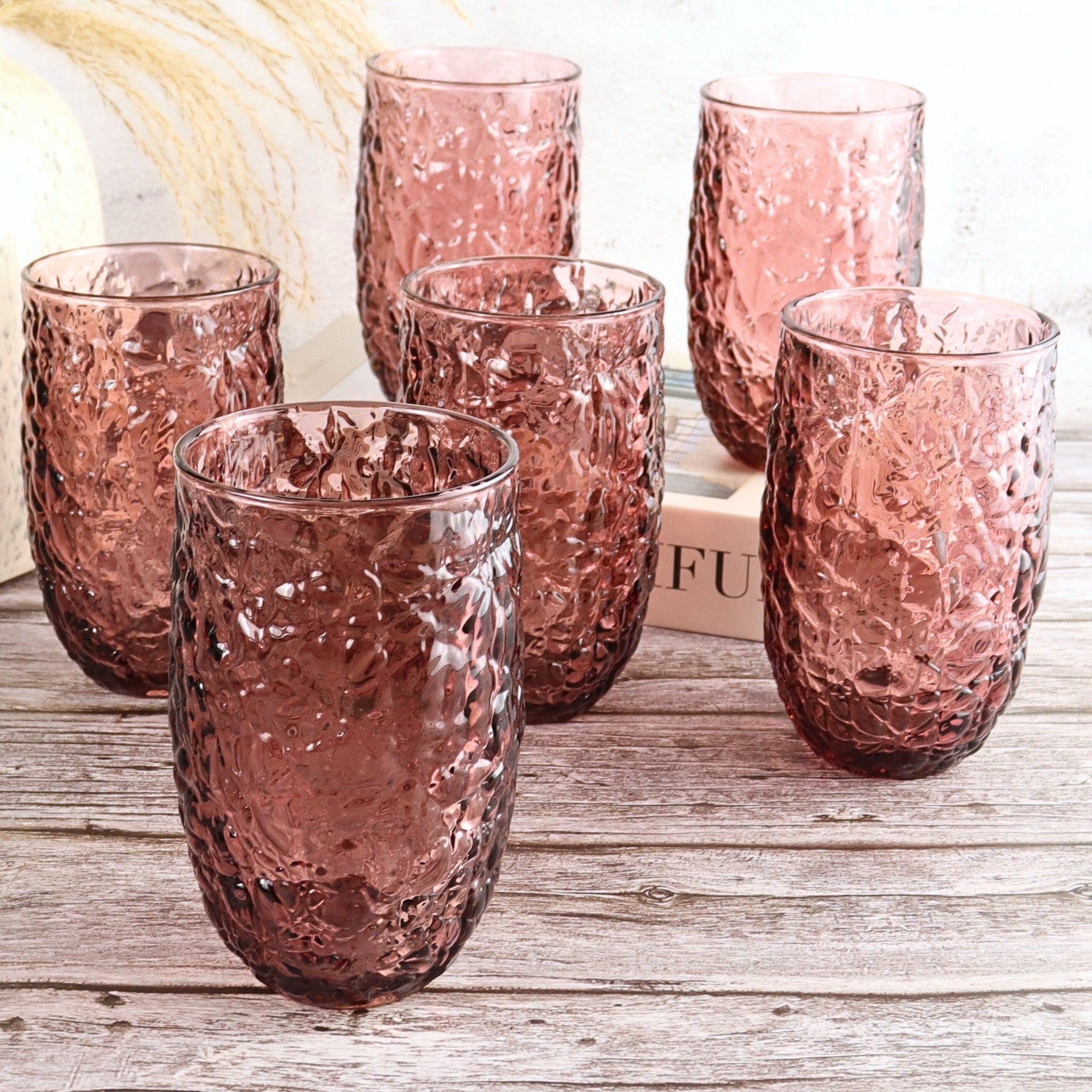 Set of six pink textured glass tumblers on a wooden surface with a blurred box in the background.